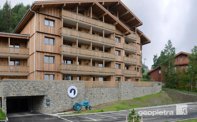 Residential building in Alpine style with wooden facades and balconies, masonry base in Geopietra manufactured stone, located in a mountainous area, with an asphalt parking lot and a blue tractor visible in the foreground.